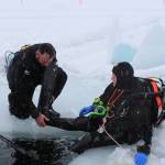 Tony Gillham, left, helps T.J. Cox with one of his flippers as Cox prepares to dive into the ice on Island Lake in Nikiski, Alaska on March 21, 2020. (Photo by Brian Mazurek/Peninsula Clarion)