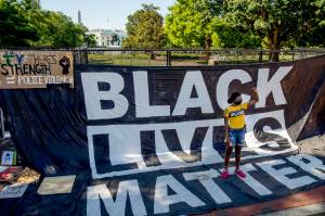 The White House is visible behind a young woman who holds her fist up as she poses for a photograph with a large banner that reads Black Lives Matter hanging on a security fence at 16th and H Street, Monday, June 8, 2020, in Washington, after days of protests over the death of George Floyd, a black man who was in police custody in Minneapolis. Floyd died after being restrained by Minneapolis police officers. (AP Photo/Andrew Harnik)
