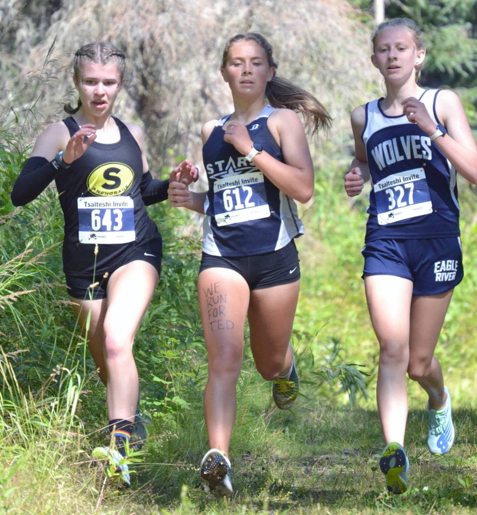 Soldotnas Jordan Strausbaugh races to 17th place in the girls varsity race at the Ted McKenney XC Invitational on Saturday, Aug. 21, 2021, at Tsalteshi Trails just outside of Soldotna, Alaska. We run for Ted is written on Strausbaughs thigh in honor of former Soldotna coach Ted McKenney. Strausbaugh runs with Souths Elizabeth Page and Eagle Rivers Ava Trembath. (Photo by Jeff Helminiak/Peninsula Clarion)