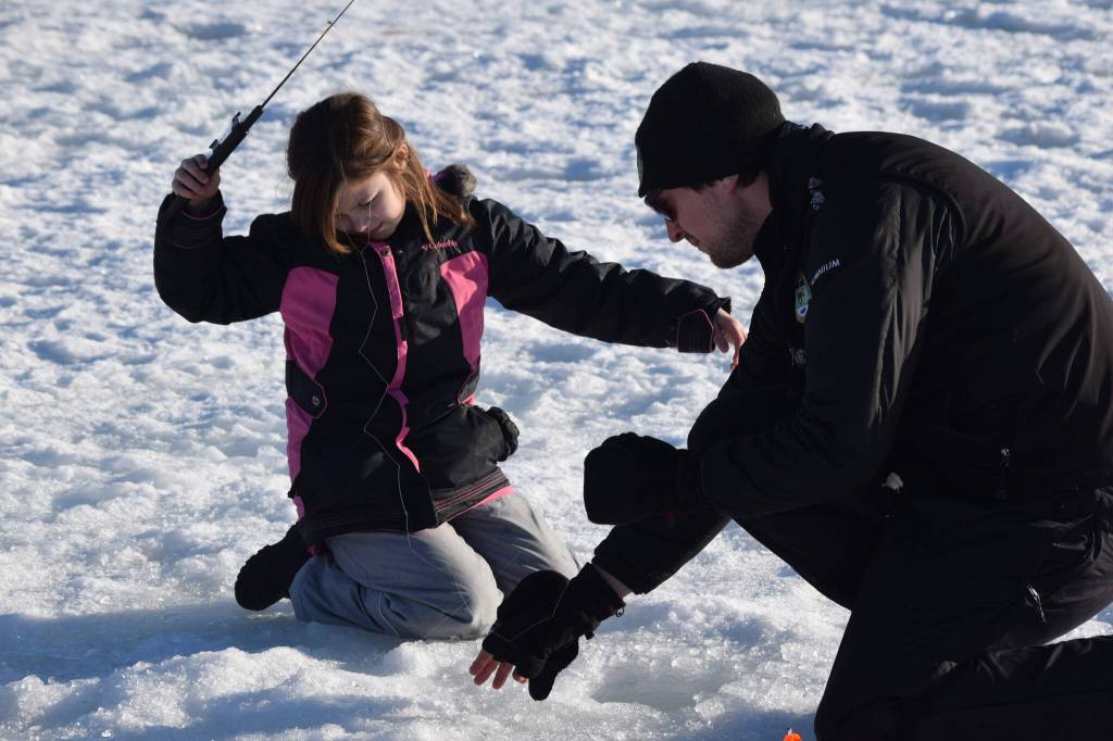 Lucas Stumpf, a fishery biologist with the Division of Sport Fish, helps Aurora Borealis Charter School third grader Teagan Jorgensen, left, with her line while she ice fishes at Sport Lake in Soldotna as part of the Alaska Department of Fish and Games Salmon in the Classroom ice fishing educational program on Wednesday, March 2, 2022, in Soldotna, Alaska. (Camille Botello/Peninsula Clarion)