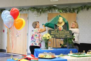Vicke Kukowski (left) and Kathy Romain (right) unveil a plaque dedicating the Kenai Senior Centers greenhouse to Bill Osborn during a ceremony on Tuesday, July 12, 2022 in Kenai, Alaska. (Ashlyn OHara/Peninsula Clarion)