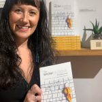 Brianna Allen poses with the book she edited, The MOMologues, at the opening of the Mother exhibit showing this month at Bunnell Street Arts Center. (Photo by Christina Whiting)