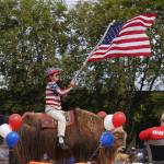 A woman waves a flag from atop a large animal as part of the Fourth of July Parade down the Kenai Spur Highway in Kenai, Alaska on Tuesday, July 4, 2023. (Jake Dye/Peninsula Clarion)