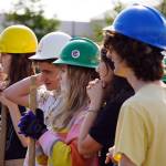 Triumvirate Theatre performers, equipped with hard hats and shovels, listen to speeches and performances at Daubenspeck Family Park in Kenai, Alaska, before a ceremonial groundbreaking for the future home of Triumvirate Theatre on Saturday, Aug. 19, 2023. (Jake Dye/Peninsula Clarion)