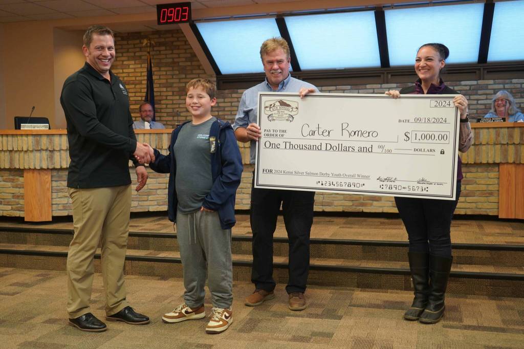 Carter Romero, center left, is awarded a $1,000 novelty check for landing the youth grand-prize salmon in the Eighth Annual Kenai Silver Salmon Derby by Kenai Mayor Brian Gabriel, a representative of Marathon Petroleums Kenai Refinery and Kenai Chamber of Commerce Executive Director Samantha Springer during a meeting of the Kenai City Council on Wednesday, Sept. 18, 2024, in Kenai, Alaska. (Jake Dye/Peninsula Clarion)