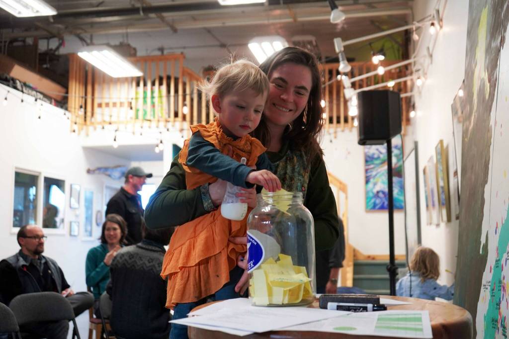 Cook Inletkeeper Central Peninsula Regional Director Kaitlin Vadla lifts up her daughter Skadi to deposit a ballot for the next local solution the organization will undertake during a Local Solutions meeting focused on salmon at the Cook Inletkeeper Community Action Studio in Soldotna, Alaska, on Thursday, March 20, 2025. (Jake Dye/Peninsula Clarion)